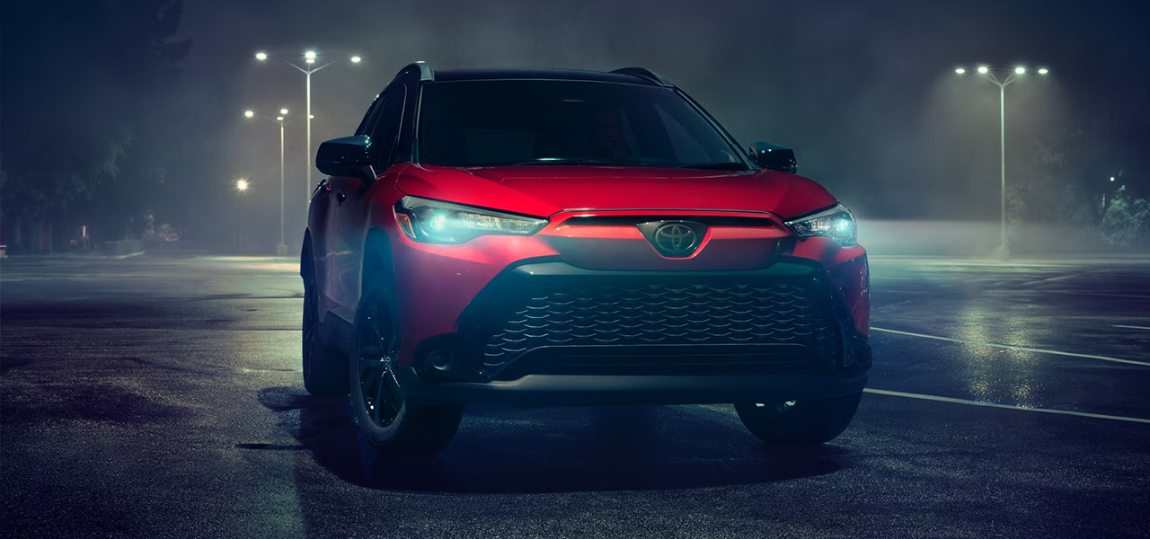 Vue 3/4 avant et latérale de la Toyota Corolla Cross 2024 dans un stationnement le soir avec les phares allumées